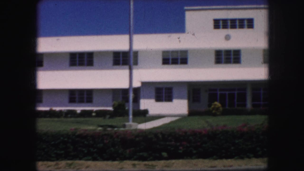 White building facade with windows, flagpole, lawn, hedge, and entrance area.