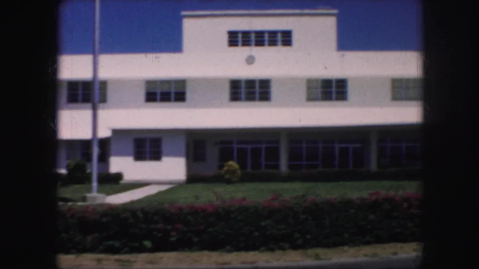 The building entrance and upper facade are seen behind a trimmed lawn, hedge row, flagpole, and flowering shrubs in 8mm footage.