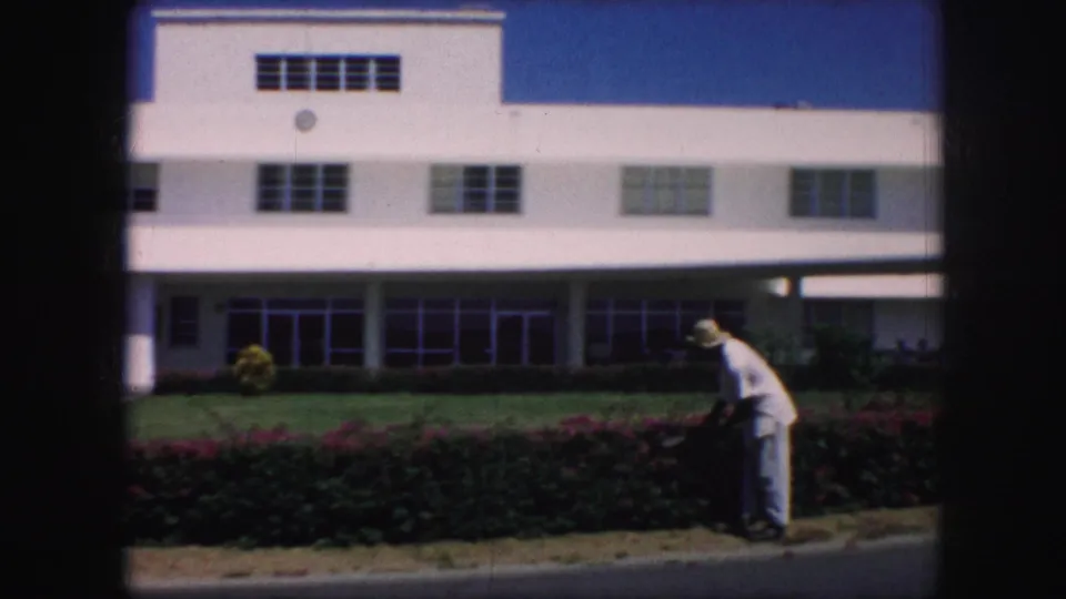 A man in a white shirt and hat trims shrubs near the curb before a white Kansas City building, filmed from a distant 8mm viewpoint.