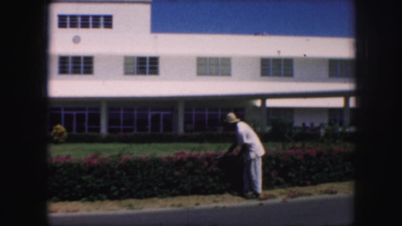 In 1959 Kansas City footage, a man in a light uniform and straw hat trims a hedge with red flowers in front of a white building.