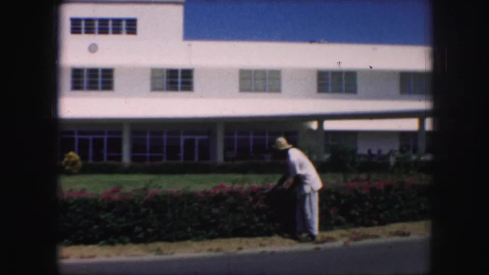 In 1959 Kansas City footage, a man in a light uniform and straw hat trims a hedge with red flowers in front of a white building.
