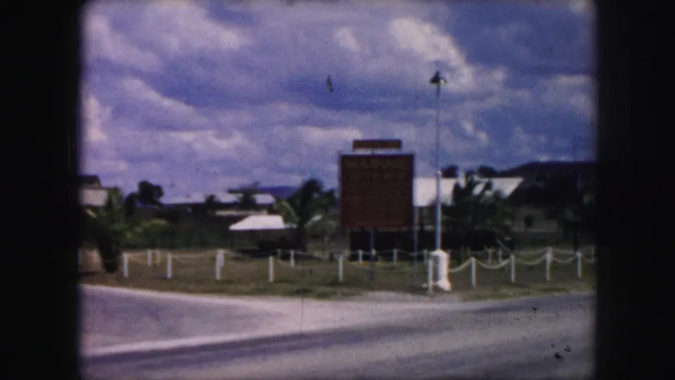 1959 8mm roadside view with a sign, flagpole, rope posts, road, and distant low buildings.