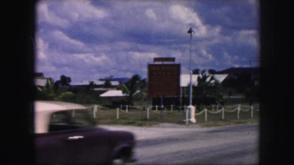 A mid-century car with a light roof passes a roadside sign in 1959 home-movie footage labeled Kansas City.