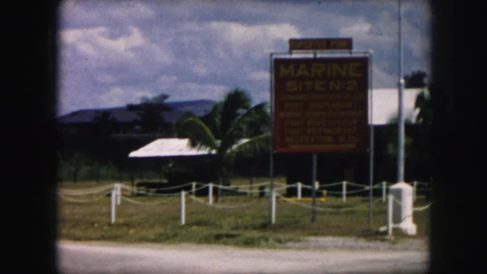 1959 8mm view of the Marine Site No. 2 sign beside a road, with palm trees, grass, and low buildings in the background.