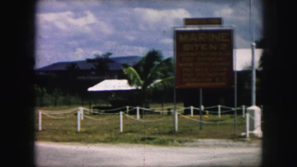 1959 8mm frame focused on the Marine Site No. 2 sign, with white boundary posts, grass, palm trees, and buildings beyond.