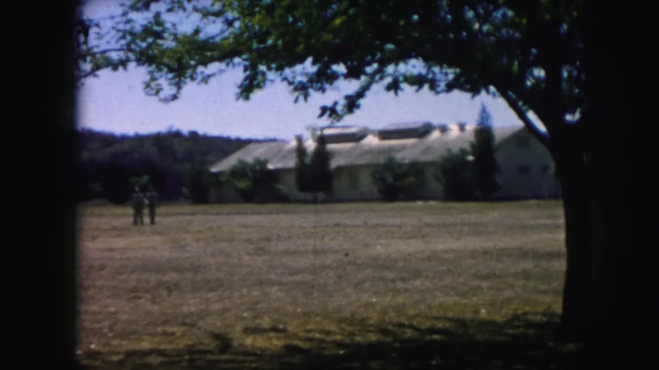 1959 Kansas City frame with two small figures crossing a dry lawn in front of a low pale building and trees.