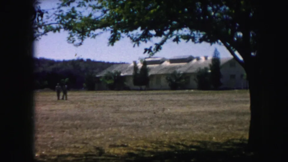1959 Kansas City home-movie view of two people on a broad lawn with a long low building and tree branches nearby.