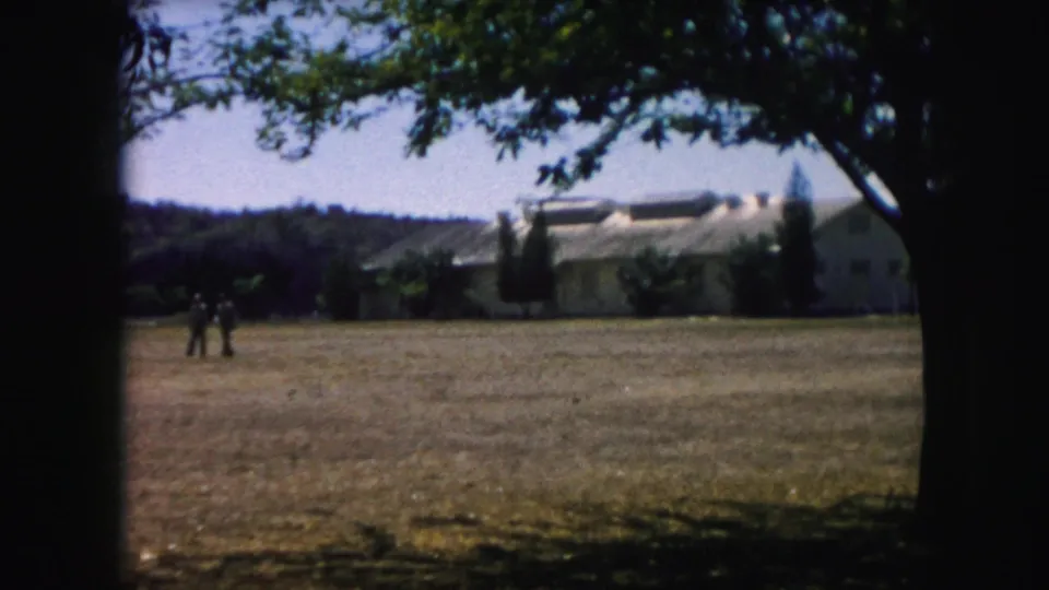 1959 Kansas City frame showing two distant figures crossing an open grassy area before a low building under tree shade.