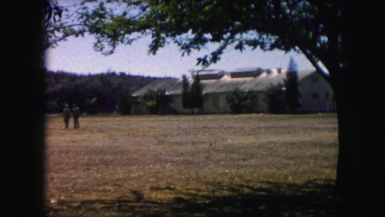 Long pale building seen across a dry lawn with trees in the foreground.