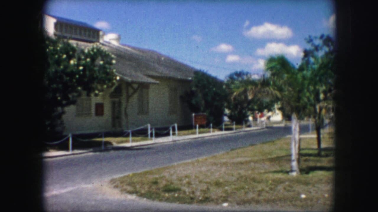 1959 Kansas City home-movie frame showing a pale building, roadside grass, palm trees, and a bright sky with scattered clouds.