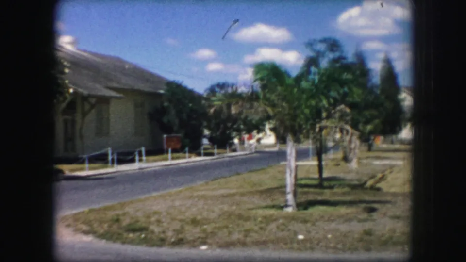 Kansas City 1959 frame with a light building set beside a paved road and lawn, framed by palms and soft 8mm edges.