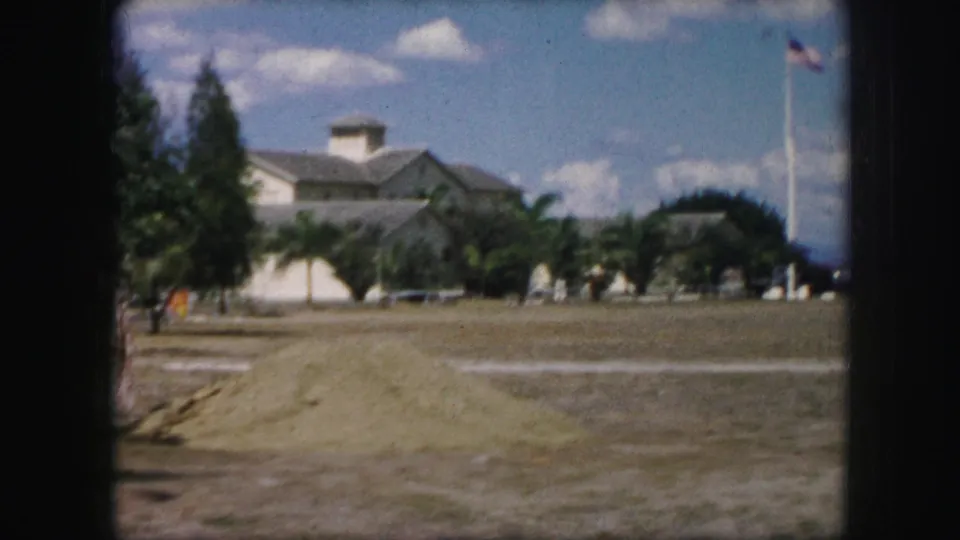 1959 Kansas City home-movie view across grass toward low buildings, palm trees, distant figures, and an American flagpole.