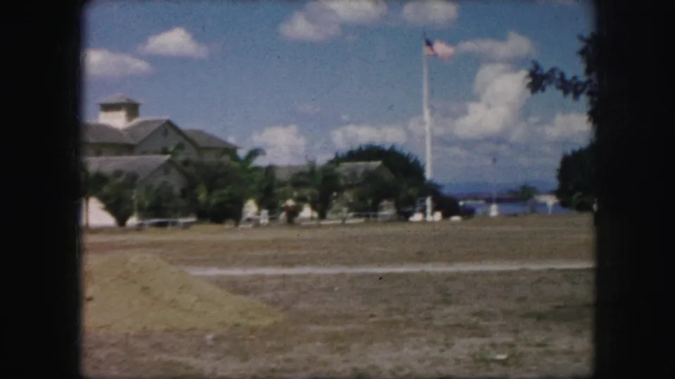 Kansas City 1959 frame looking across open grounds toward palms, a low building, distant people, and an American flag on a tall pole.