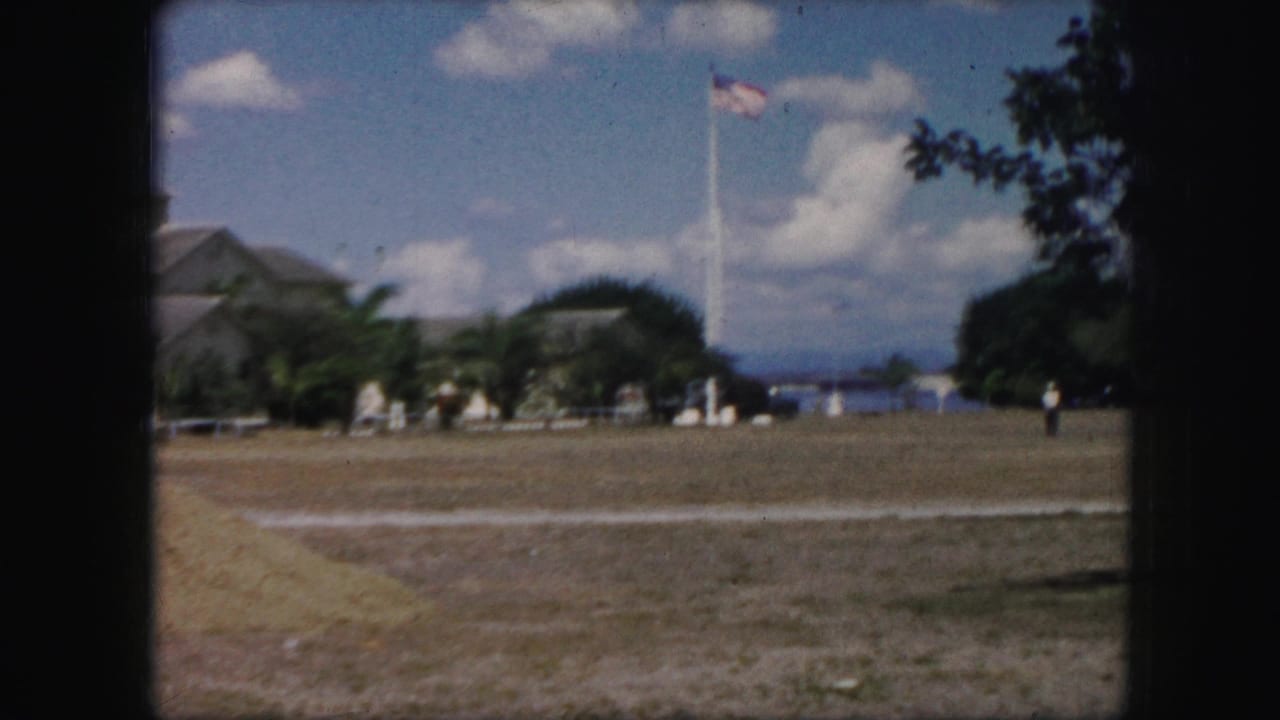 1959 8mm frame showing an open public field with a tall American flagpole, a distant walking figure, trees, clouds, and water visible in the background.