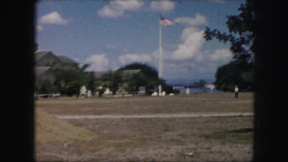 1959 8mm frame showing an open public field with a tall American flagpole, a distant walking figure, trees, clouds, and water visible in the background.