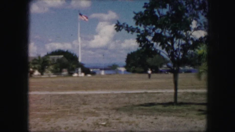 1959 8mm frame of a broad field with a raised American flag, scattered trees, a distant person, and water beyond the grounds.