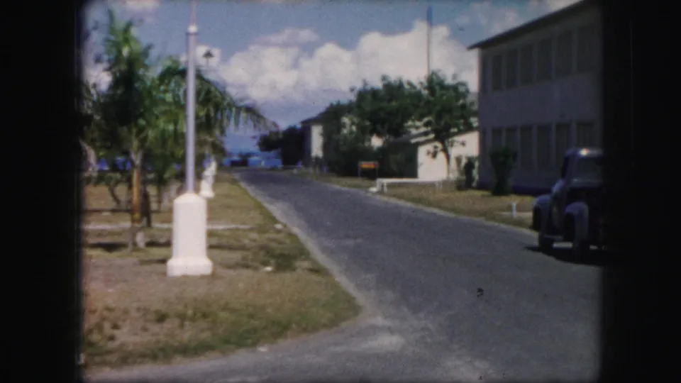 1959 8mm frame of a paved roadside scene with a white lamppost, modest buildings, trees, distant water, and a parked vintage truck or car.