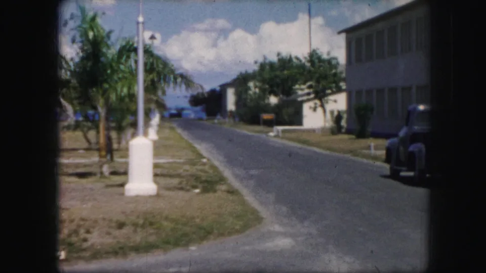 1959 8mm frame showing a quiet palm-lined street with low buildings, a roadside post, and a dark parked car at the right edge.
