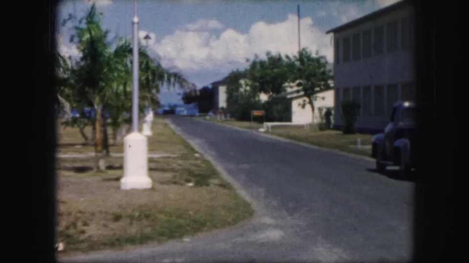 1959 8mm frame of a quiet road bordered by palms and low buildings, with a dark car partly visible along the right side.