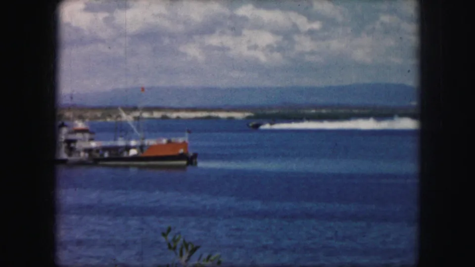 1959 8mm frame showing a bay with small boats in the foreground, a speedboat wake across the water, distant hills, and a clouded sky.