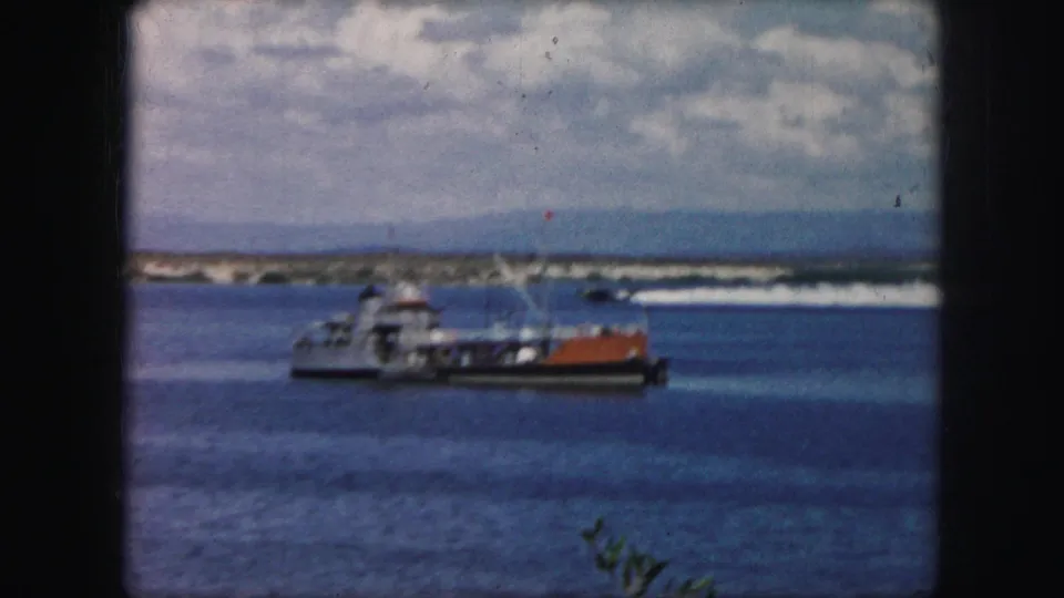 1959 8mm frame of small boats on a bright bay with a white speedboat wake, distant landforms, and a partly cloudy sky.