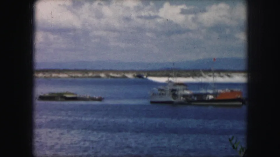 1959 8mm frame showing a ferry or workboat and a low barge on open water, with shoreline and clouds in the background.