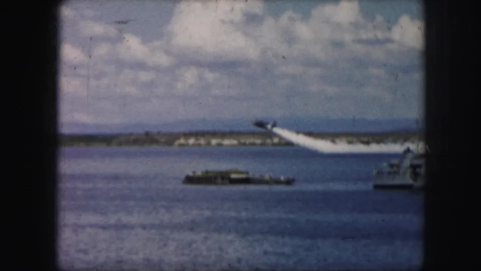 1959 8mm frame showing a small plane rising over the water while boats sit below and a white smoke trail follows behind it.
