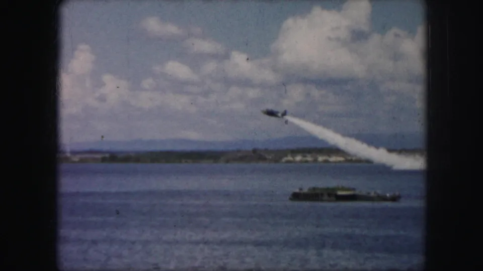 1959 8mm frame showing a small plane banking or climbing over water, leaving a white smoke trail above a low barge.