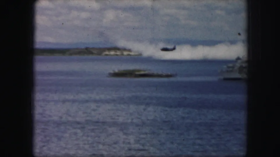 1950s 8mm frame of a dark aircraft passing over open water, seen through a dark vertical frame with distant shore and boats.