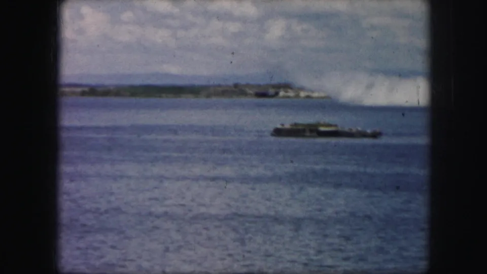 1950s 8mm frame showing a dark low aircraft near a white smoke trail above water, with a boat and distant shoreline visible.