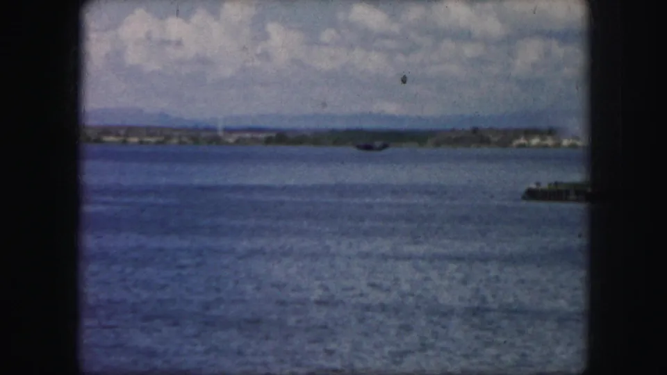 1950s 8mm frame of a small dark aircraft in the distance over water, with boats, shoreline, clouds, and window-frame edges.