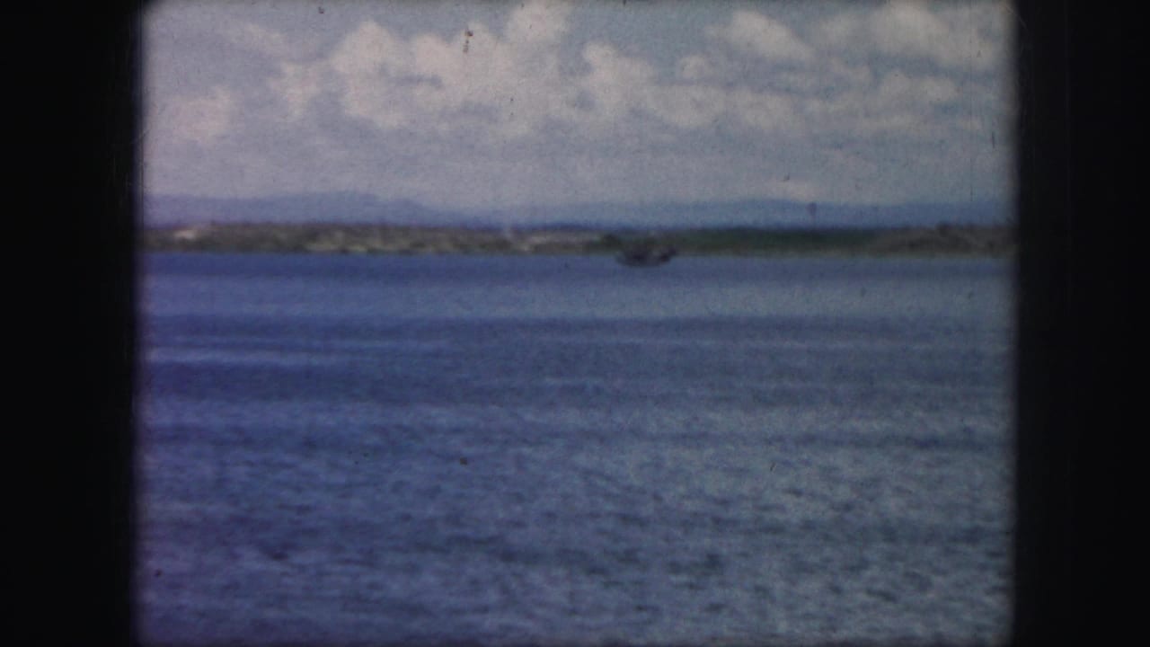 1959 8mm frame from Kansas City, Missouri metadata showing open blue water, a far shoreline, and clouds before the aircraft becomes clear.