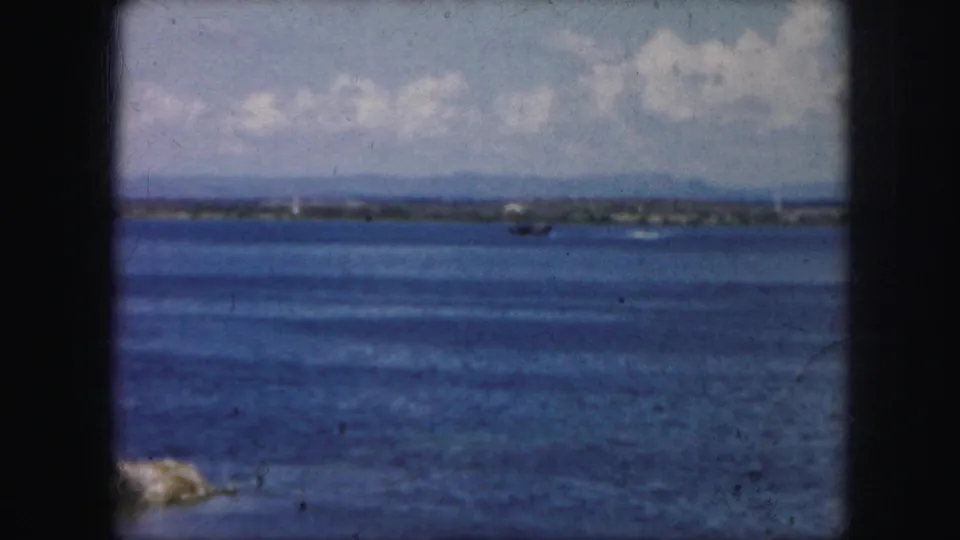 Kansas City-area 1959 home-movie frame showing lake water, distant land, and a small wake or aircraft trace near the shoreline.