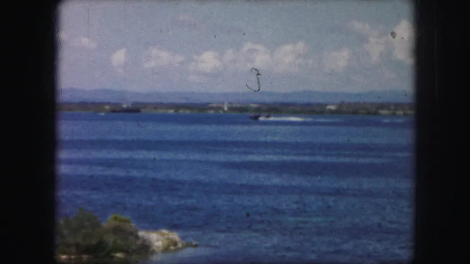 1959 8mm frame showing a small aircraft or seaplane on or just above the water, leaving a bright wake across the lake.