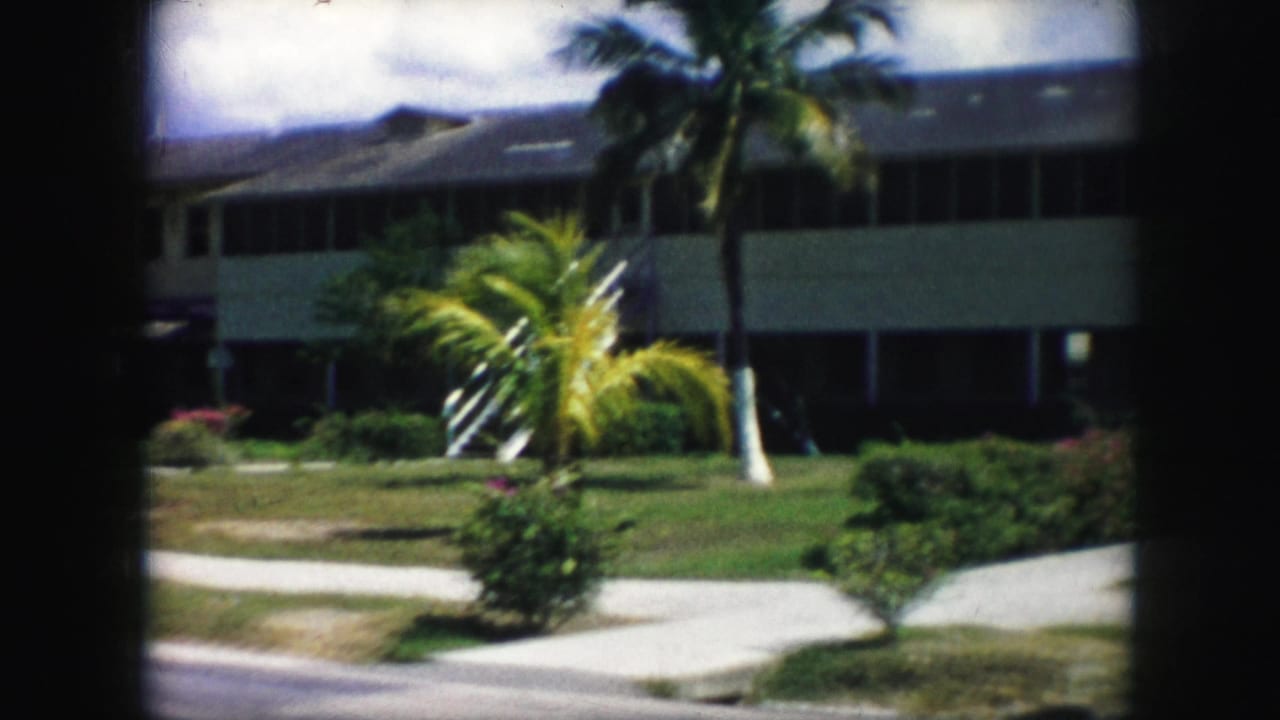 1959 home-movie frame of a two-story modern building behind palms, lawns, and concrete walkways, with dark film-scan borders.