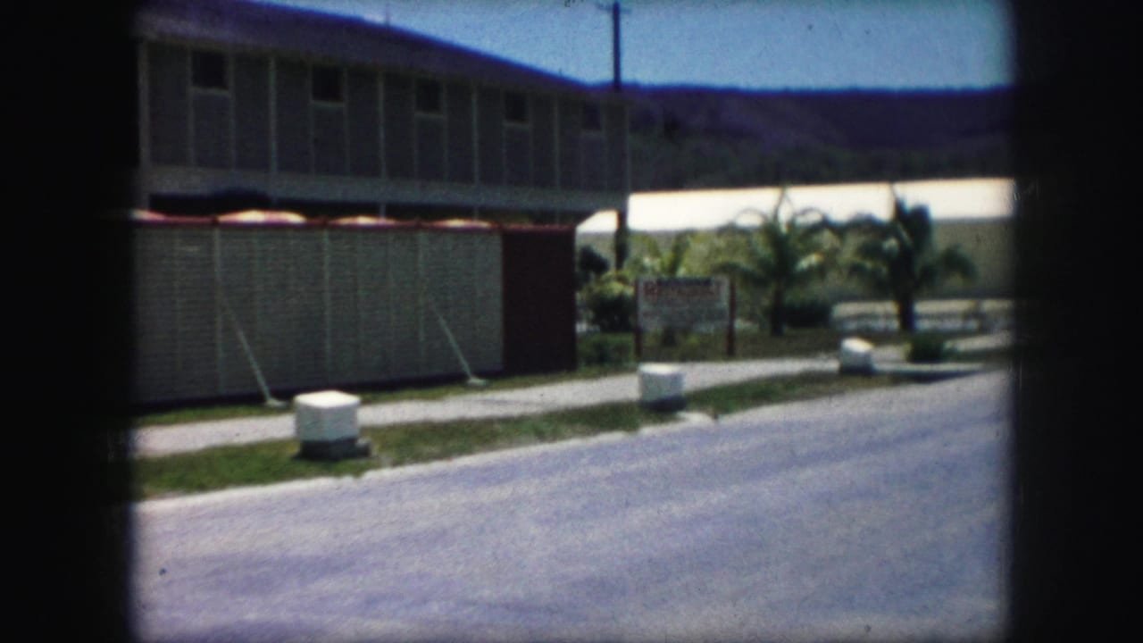 Roadside frame showing a modern exterior with a screened or fenced section, small sign, palm plantings, parked car, and hills beyond.