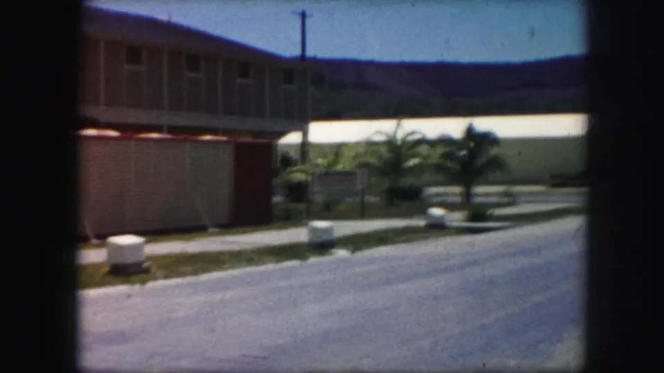 Soft 8mm frame of a roadside modern building with a sign, palm landscaping, low walls, utility pole, and distant hillside.