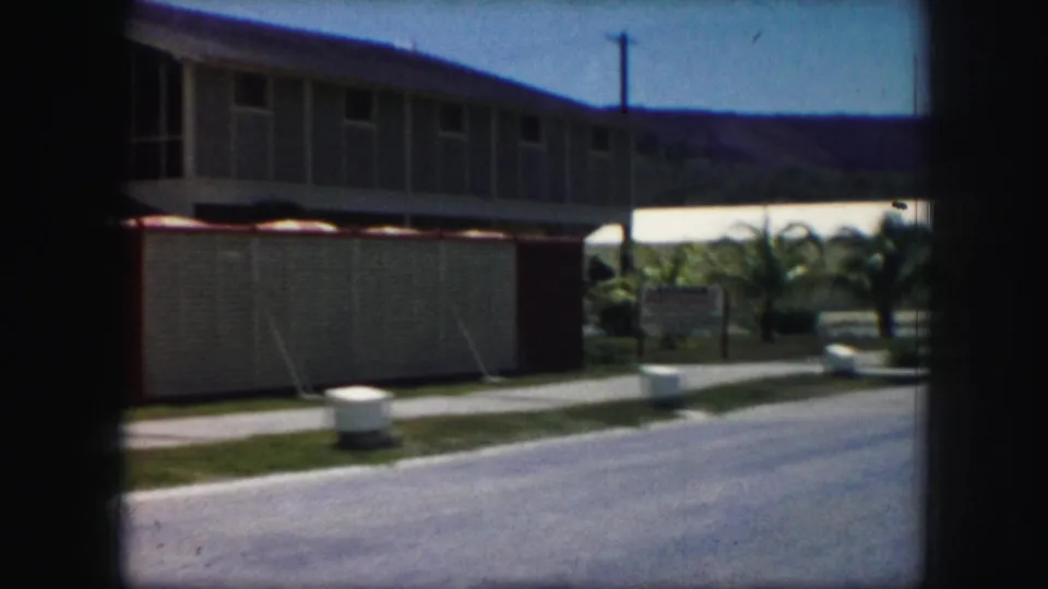 Final frame holds on a modern roadside building with a screened section, small sign, palm landscaping, parked car, and distant hills.