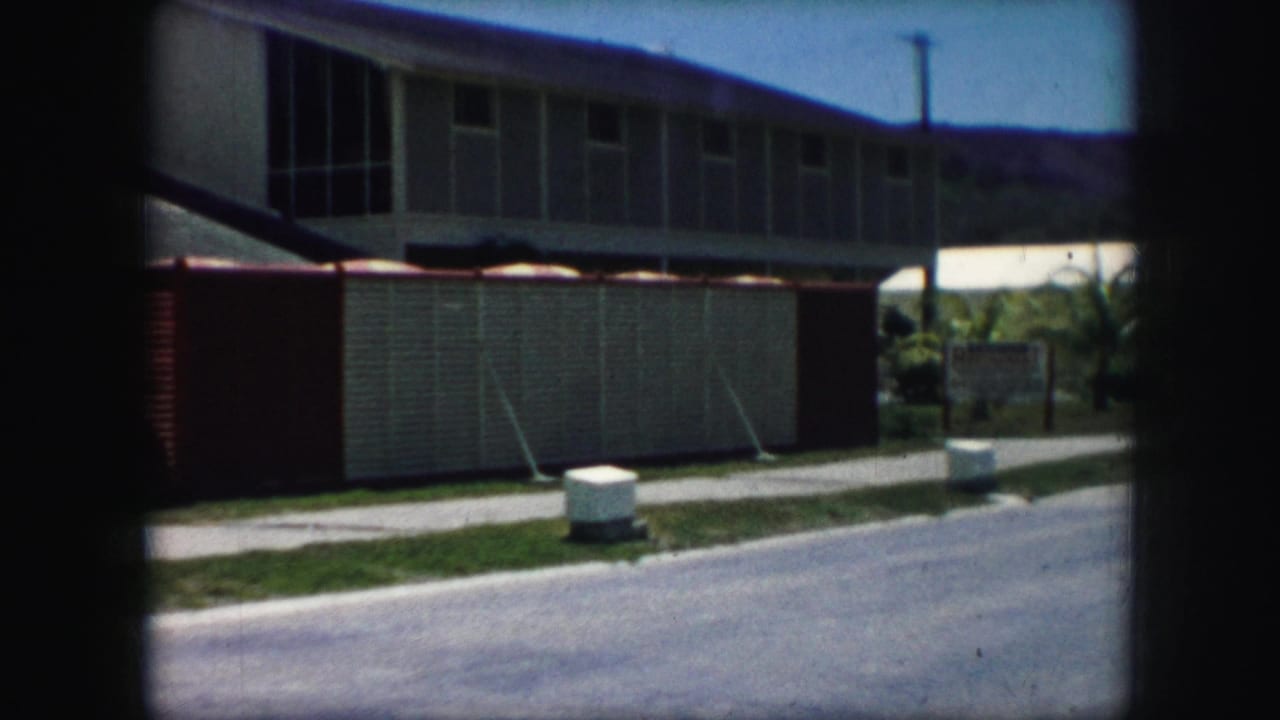1959 Kansas City home-movie frame showing a sloped-roof house, sidewalk, curb, and block wall across a residential street.