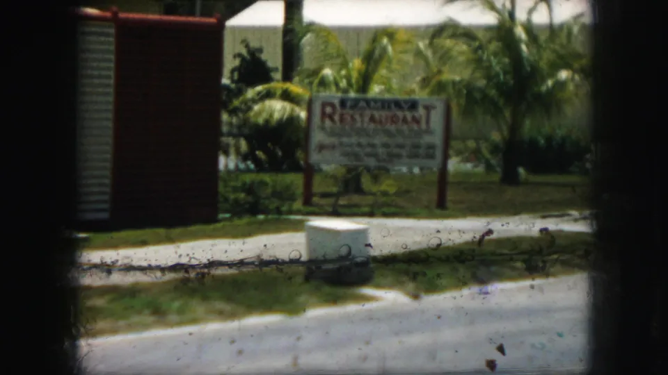1959 Kansas City home-movie frame with a roadside restaurant sign partly blurred by motion, bordered by grass, curb, palms, and film artifacts.