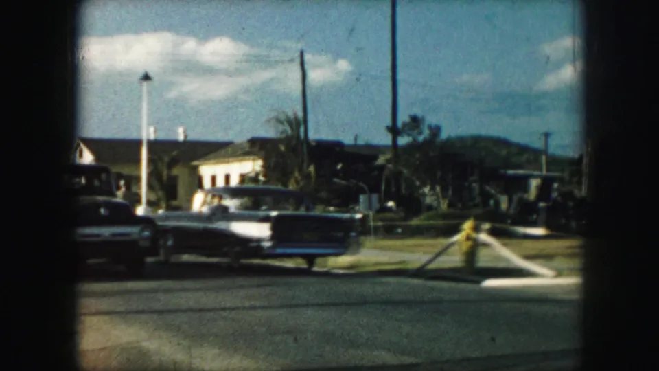 1959 Kansas City street view showing classic cars driving past low buildings, utility poles, a bright sky, and a roadside barrier.