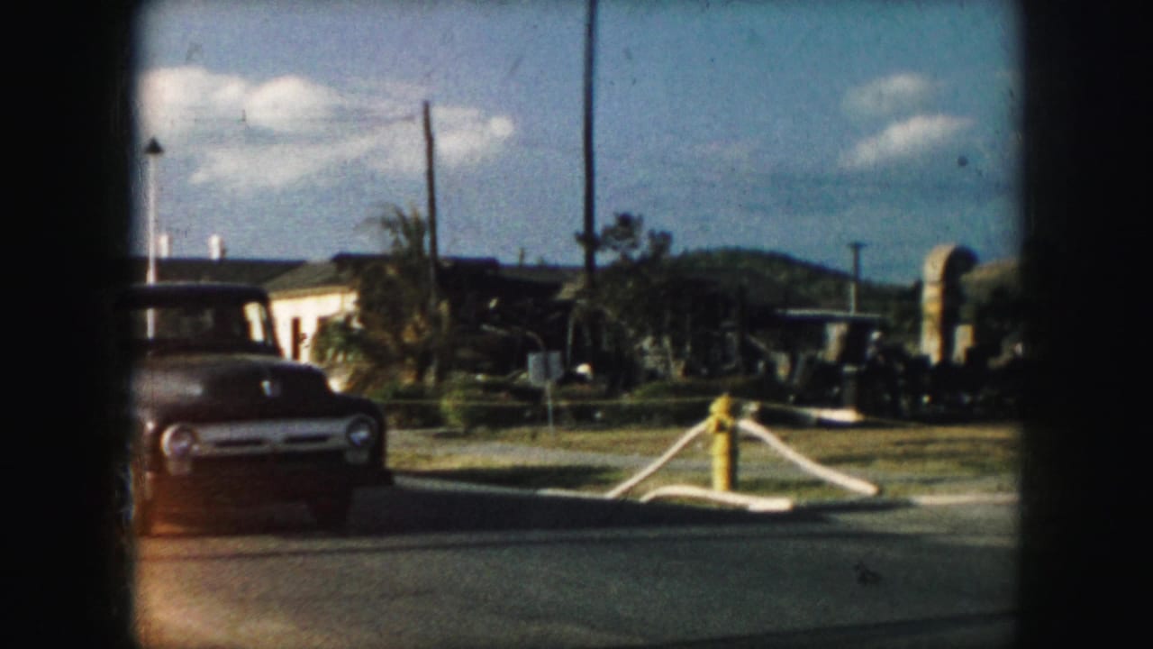 Older car near a yellow fire hydrant on a sunny street with low buildings behind it.