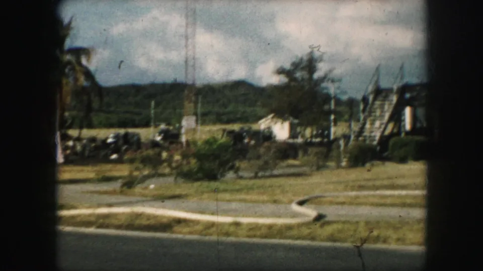 1959 8mm frame showing a curving paved path, grass, parked vehicles, hillside, and a structure with exterior stairs.