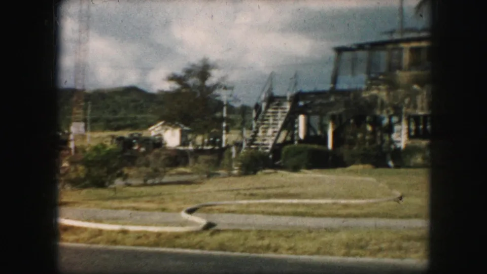 Late frame from a 1959 8mm roadside view, with a winding path, grass, distant hills, parked vehicles, and exterior stairs on a raised structure.