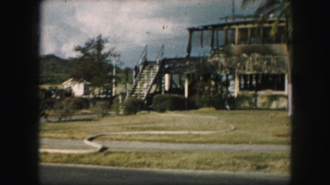 1959 8mm frame showing a large house with exterior stairs, a broad lawn, shrubs, and a road edge in the foreground.