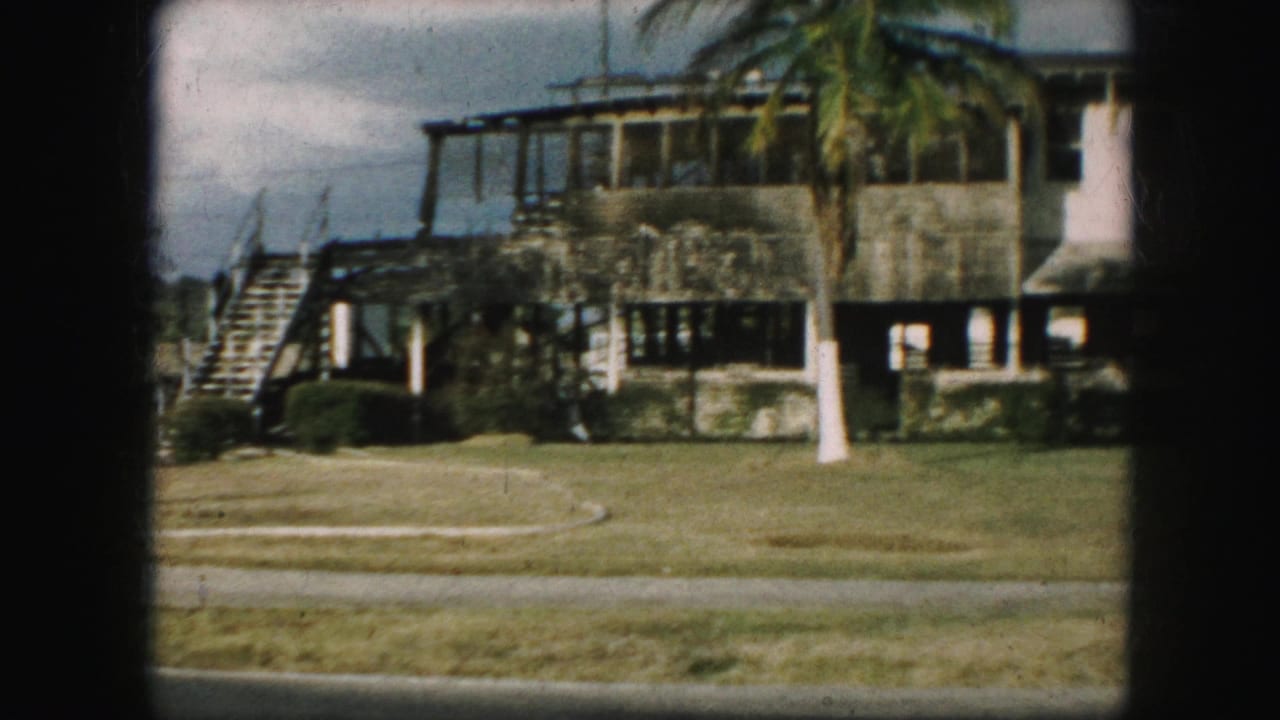 1959 8mm view of a large residential facade with stairs, lawn, palm-like landscaping, shrubs, and visible film borders.