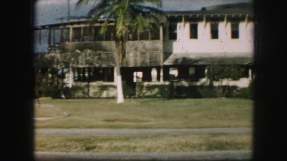 1959 8mm frame with the house closer in view, showing lawn, palm trunk, upper windows, shaded porch area, and soft film grain.