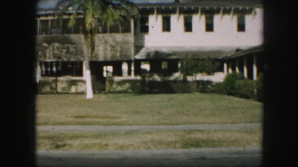 1959 8mm frame showing a large house beyond a lawn, with a palm-like tree, shrubs, porch openings, and roadside foreground.