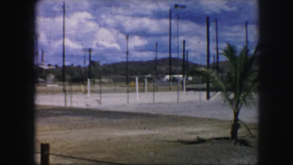1959 Kansas City 8mm frame showing a palm tree at the edge of a broad paved lot with utility poles, fencing, low hills, and dramatic clouds.