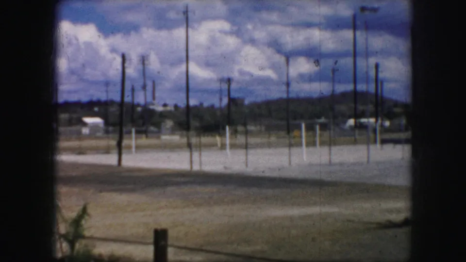 Kansas City 1959 8mm view of a broad empty lot with utility poles, fence posts, low hills, heavy clouds, and a partial palm frond in the foreground.